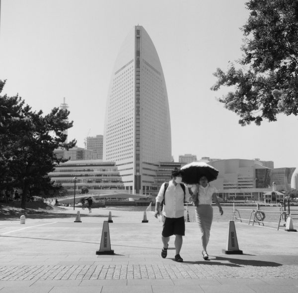 Two passersby walk along a Yokohama port at midday, shielding themselves from the sun with an umbrella. Most pedestrians wore light-colored, loose-fitting clothing to reflect sunlight, though shorts were rare.
